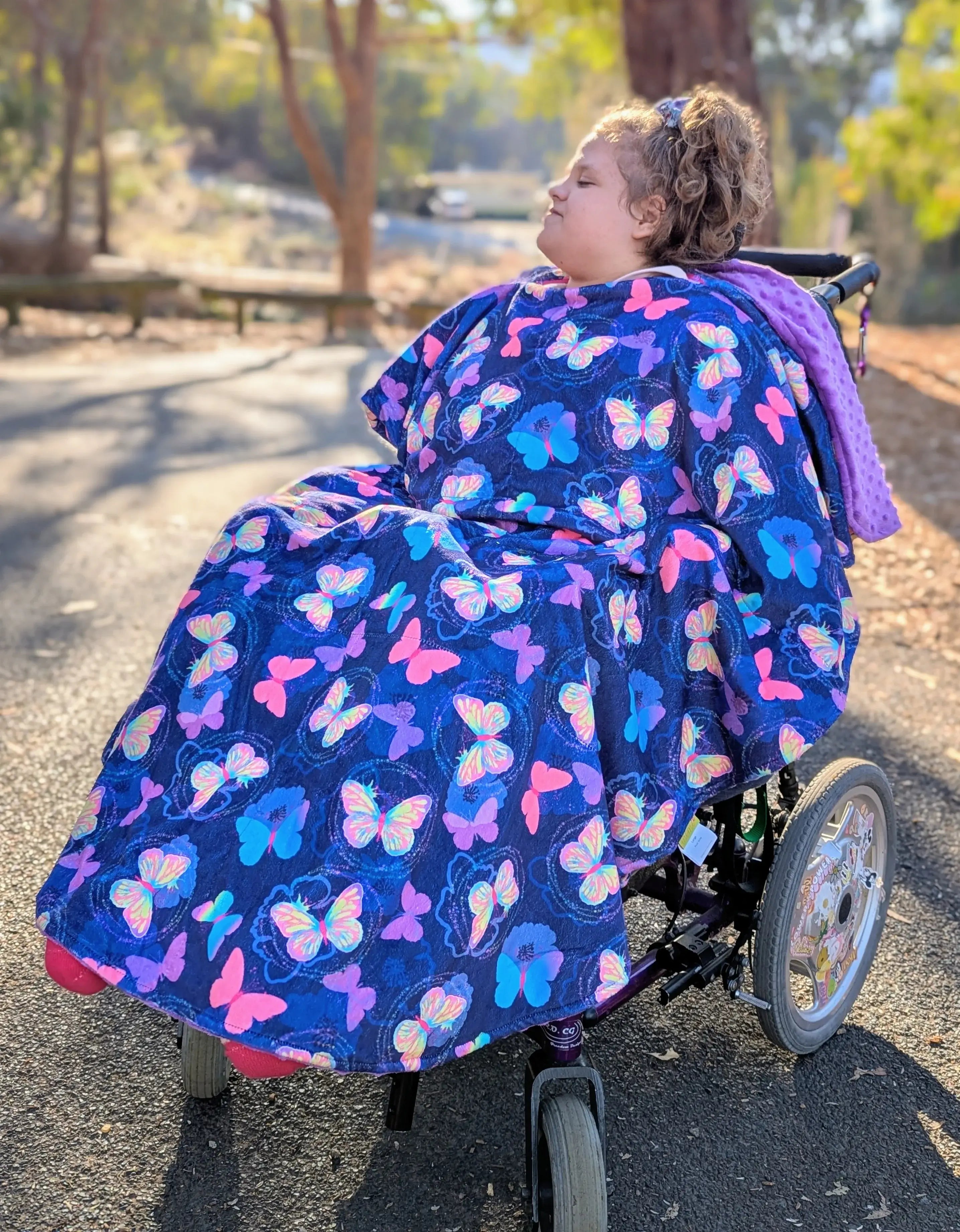 Child in a wheelchair covered with a colorful butterfly-patterned blanket outdoors.